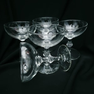 May include: A set of four clear glass coupe glasses with wide, shallow bowls and short, decorative stems. Three glasses stand upright, while one glass is positioned horizontally in the foreground, showcasing the circular base and stem design.