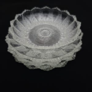 May include: A stack of three clear glass bowls featuring a scalloped edge and a detailed pressed pattern. The design includes a central starburst motif surrounded by radiating geometric shapes and textured panels that catch the light.