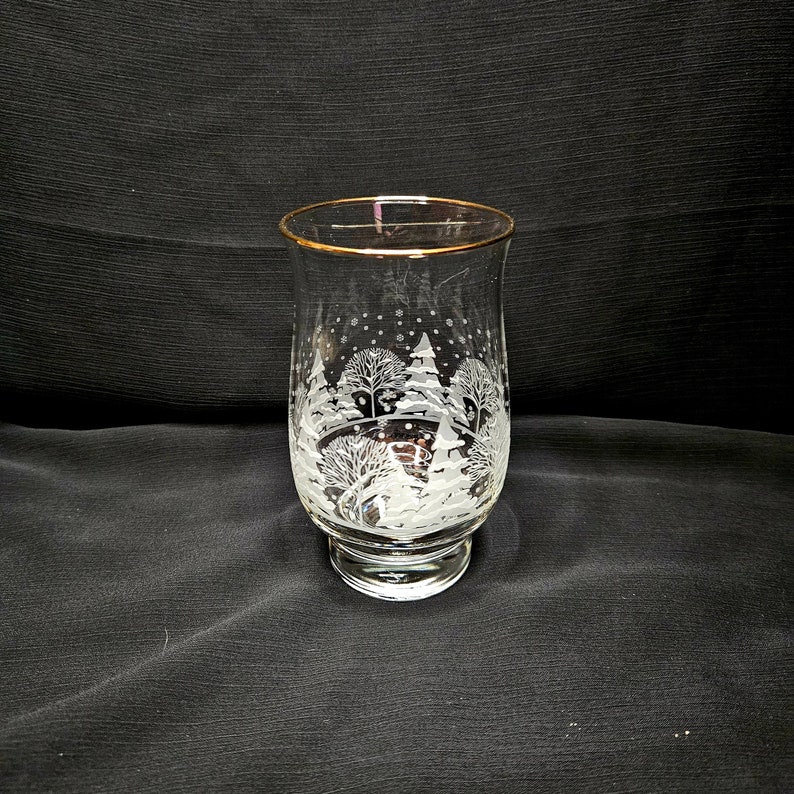 May include: A clear glass vase with a gold rim and a winter scene etched into the side. The scene features trees, snow, and snowflakes.