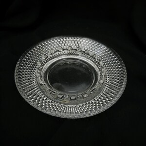 May include: A clear glass plate featuring a textured diamond pattern across the rim and a smooth, circular center. The inner edge of the plate is decorated with a series of small, raised circular indentations, creating a detailed border.