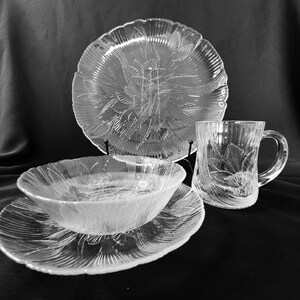 May include: A set of clear glass dinnerware with a floral pattern. The set includes a large plate, a smaller plate, a bowl, and a mug.