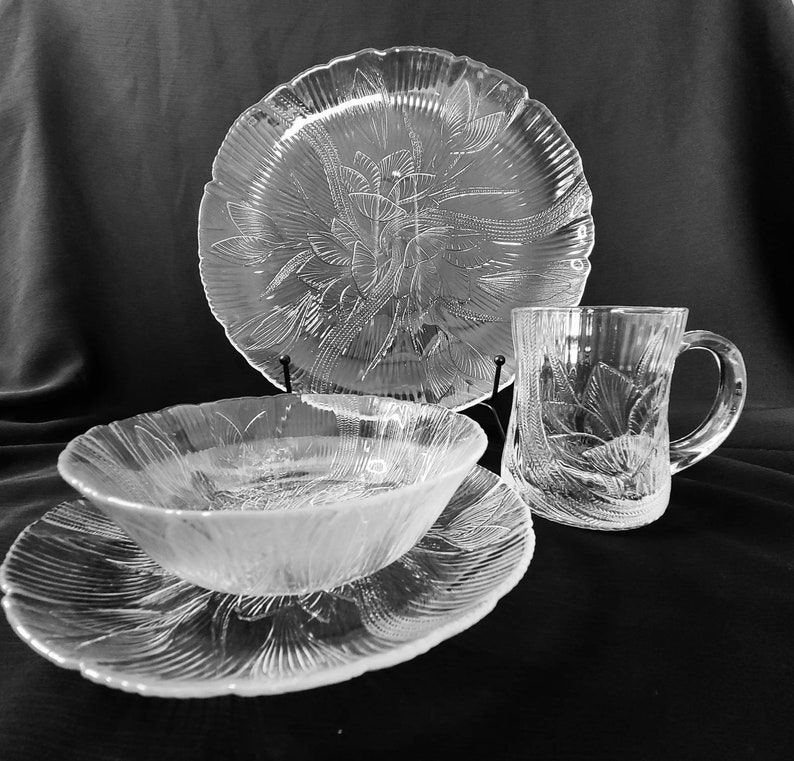 May include: A set of clear glass dinnerware with a floral pattern. The set includes a large plate, a smaller plate, a bowl, and a mug.
