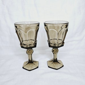 May include: Two clear glass wine goblets with a dark brown tint and a faceted design. The goblets have a round bowl and a tall stem with a wide base.