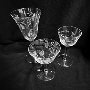May include: Three clear, stemmed glasses with etched floral and berry designs. The glasses vary in shape and size, with one tall, flared glass and two shorter, rounded glasses. The etched details add a delicate touch to the glassware.