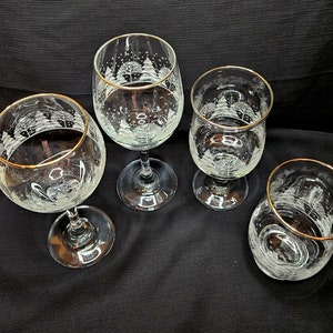May include: Four clear glass wine glasses with gold rims. Each glass has a white etched design of a winter scene with trees and snow.