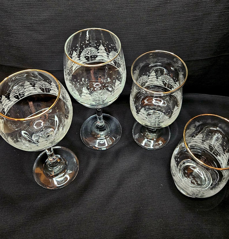 May include: Four clear glass wine glasses with gold rims. Each glass has a white etched design of a winter scene with trees and snow.