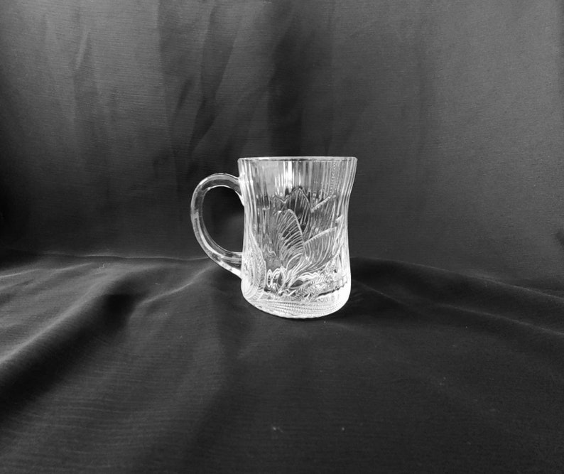 May include: A clear glass mug with a decorative etched design. The mug has a handle and a wide base.