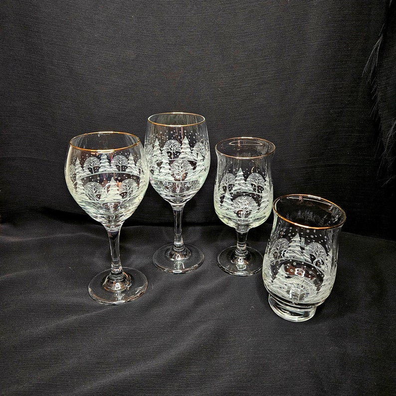 May include: Four clear glass wine glasses with gold rims. Each glass features an etched design of a winter scene with trees and snow.