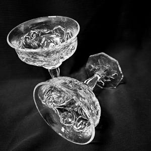 May include: Two clear, stemmed champagne glasses with ornate floral patterns. The glasses are made of glass and are positioned at an angle against a dark background. The stems have a unique design.