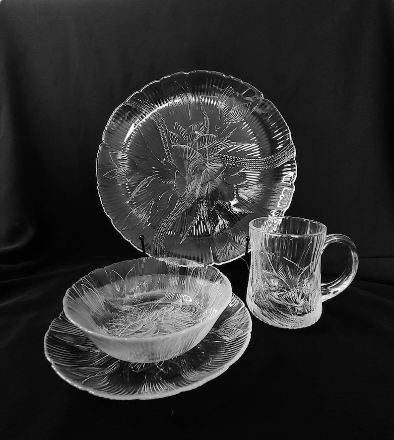 May include: A set of clear glass dishes with a floral pattern. The set includes a large plate, a smaller plate, a bowl, and a mug.