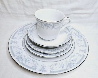 May include: A set of four white porcelain plates with a blue and silver floral pattern. The plates are stacked on top of each other, with a matching teacup and saucer on top.
