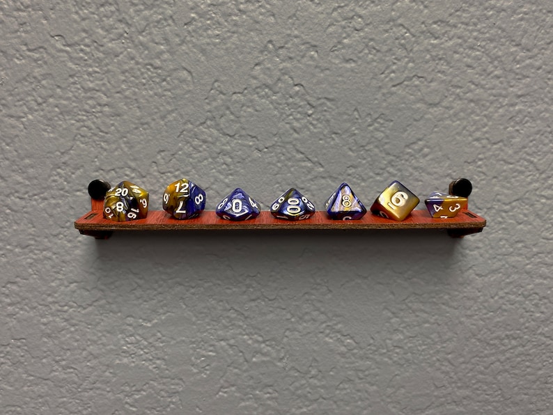 May include: A wooden shelf with seven dice, including a 20-sided die, a 12-sided die, a 4-sided die, a 6-sided die, a 8-sided die, a 10-sided die, and a 4-sided die. The dice are various colours, including gold, blue, and black.