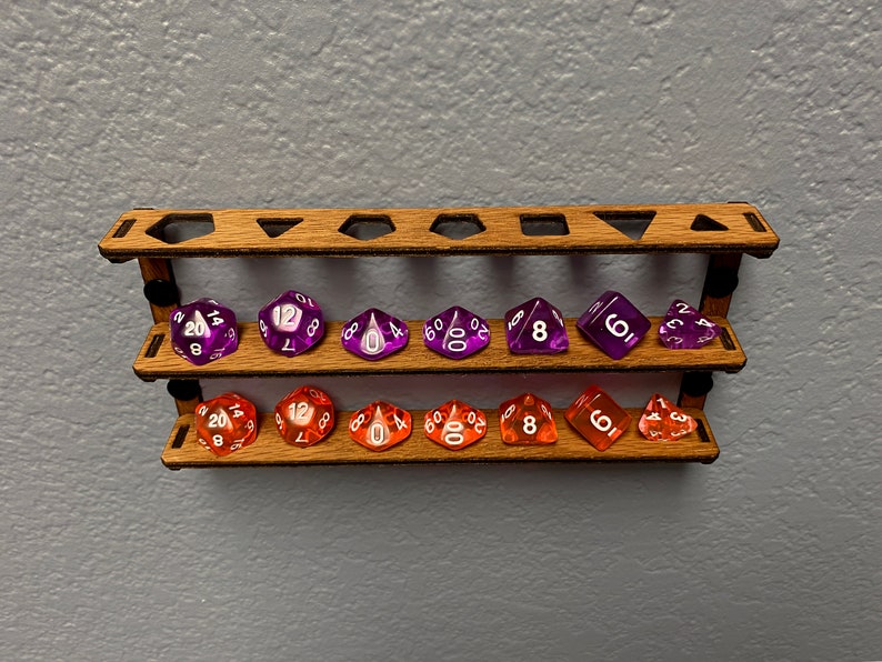May include: A wooden dice holder with two shelves, each holding seven dice. The top shelf has purple dice with white numbers, and the bottom shelf has orange dice with white numbers.
