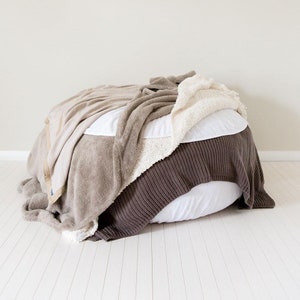 May include: A white round beanbag chair covered with a brown knitted blanket, a beige fuzzy blanket, and a white fluffy blanket.
