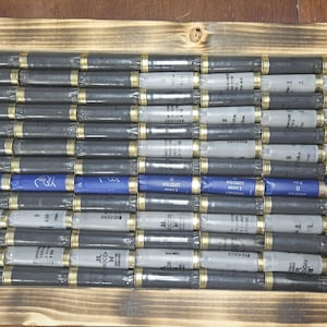May include: A wooden framed art piece featuring a design made from shotgun shells. The shells are arranged to resemble a flag, with rows of black, gray, and blue shells. The frame has a dark, burnt wood finish.