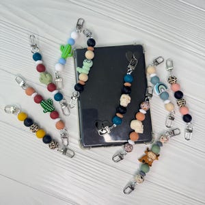 Puede incluir: Una colección de colgantes para teléfono con cuentas de varios colores y diseños. Los colgantes presentan una variedad de formas, incluyendo cactus, calaveras e impresiones de animales. Cada colgante tiene un cierre plateado para una fácil fijación.