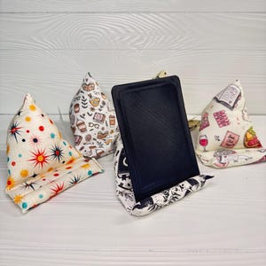 Puede incluir: Cuatro soportes de tela tipo cojín para tabletas o libros, con varios patrones que incluyen estrellas coloridas, tazas de café e ilustraciones de libros. Un soporte sostiene una tableta rectangular negra en posición vertical.