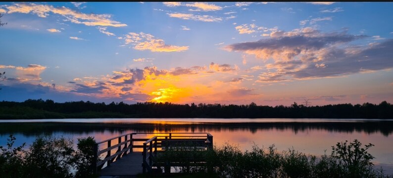May include: A scenic landscape photograph of a lake at sunset. The sky is a mix of blue, orange, and yellow hues, with fluffy clouds. A wooden pier extends into the calm water, reflecting the colorful sky. Trees line the horizon.