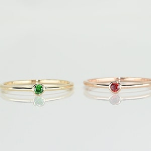 May include: Two simple gold rings with a single gemstone. The ring on the left has a green gemstone, and the ring on the right has a red gemstone.