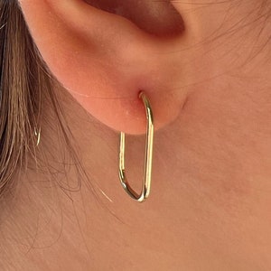 May include: Gold hoop earrings with a rectangular shape. The earrings are worn in a person's ear.