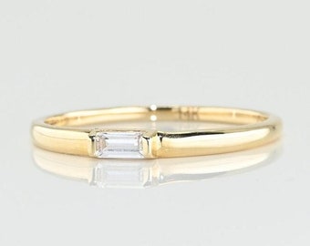 May include: A delicate gold band ring with a single, small, rectangular-shaped diamond set in the center.