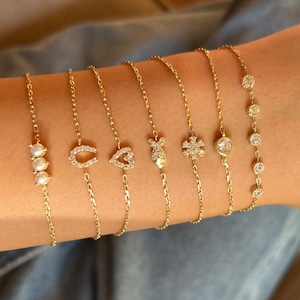 May include: A set of seven gold chain bracelets with different charms, including a horseshoe, a heart, a fish, a snowflake, a single round stone, and a row of three pearls.