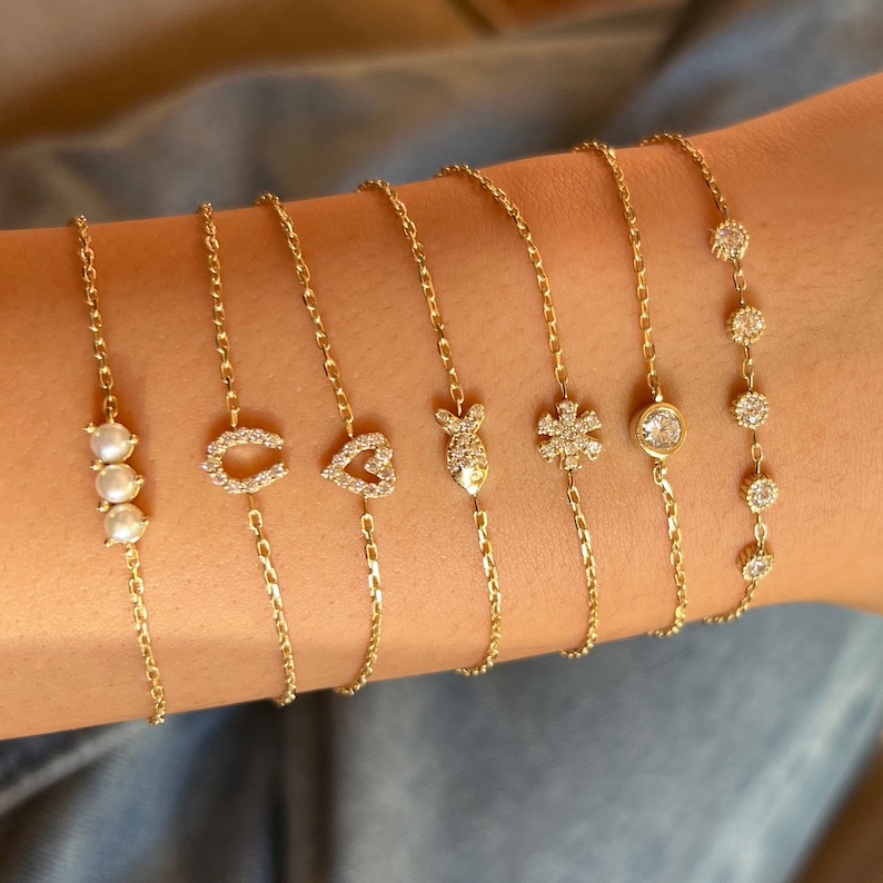 May include: A set of seven gold chain bracelets with different charms, including a horseshoe, a heart, a fish, a snowflake, a single round stone, and a row of three pearls.