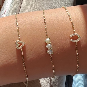 May include: Three gold chain bracelets with different charms. One bracelet has a heart charm with small clear stones, another has three pearl charms, and the third has a horseshoe charm with small clear stones.
