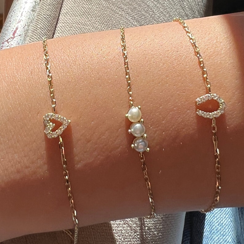 May include: Three gold chain bracelets with different charms. One bracelet has a heart charm with small clear stones, another has three pearl charms, and the third has a horseshoe charm with small clear stones.