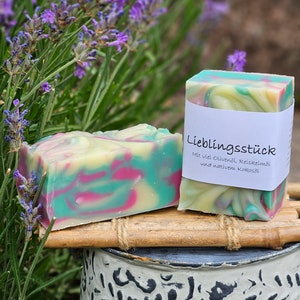 Könnte beinhalten: Zwei handgemachte Seifen mit grünen, rosa und weißen Wirbeln. Die Seife rechts hat ein Etikett mit der Aufschrift "Lieblingsstück" mit dem Text "Mit viel Olivenöl, Reiskeimöl und nativem Kokosöl".
