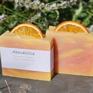 Könnte beinhalten: Zwei handgemachte Seifenstücke mit einer getrockneten Orangenscheibe darauf. Die Seife ist hellgelb mit einem rosa Wirbel. Das Etikett auf der Seife lautet "Abendstlle" mit dem Text "Mit nativem Olivenöl, Kokosöl und Reiskeimöl".