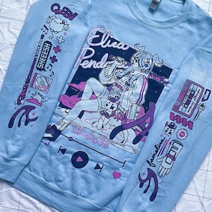 Könnte beinhalten: Hellblauer Pullover mit einem Grafikdruck eines Comic-Charakters mit rosa Haaren und einem rosa-weißen Outfit. Der Charakter hält ein Mikrofon und der Text "Elica Pend" befindet sich über dem Charakter. Der Pullover hat auch andere grafische Elemente, darunter ein Kassettenband, einen Musikplayer und den Text "Trial & Error".