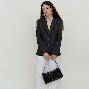 Könnte beinhalten: Ein dunkelgrauer Nadelstreifen-Blazer mit Zwei-Knopf-Verschluss, kombiniert mit einer weißen Hose mit weitem Bein und einer schwarzen Handtasche. Das Outfit wird durch Perlenschmuck und schwarze Sandalen ergänzt.