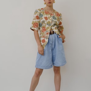 Könnte beinhalten: Eine Vintage-Bluse mit floralem Muster, kurzen Ärmeln und goldenen Knöpfen, kombiniert mit hoch taillierten hellblauen Jeansshorts. Das Outfit wird mit einem braunen Gürtel und schwarzen Riemchen-High Heels ergänzt. Der Gesamtstil ist retro und schick.