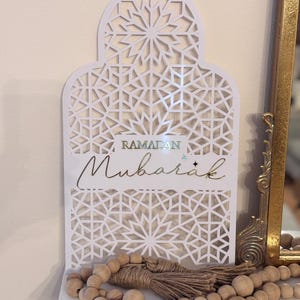 May include: White laser-cut Ramadan Mubarak sign with geometric patterns. The sign features the words "Ramadan Mubarak" in gold script. A wooden bead garland and tassel are in front of the sign.
