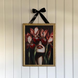 May include: A framed print of a woman's face with red and white iris flowers covering her eyes. The flowers are in focus, and the woman's face is blurred. The frame is gold and ornate.