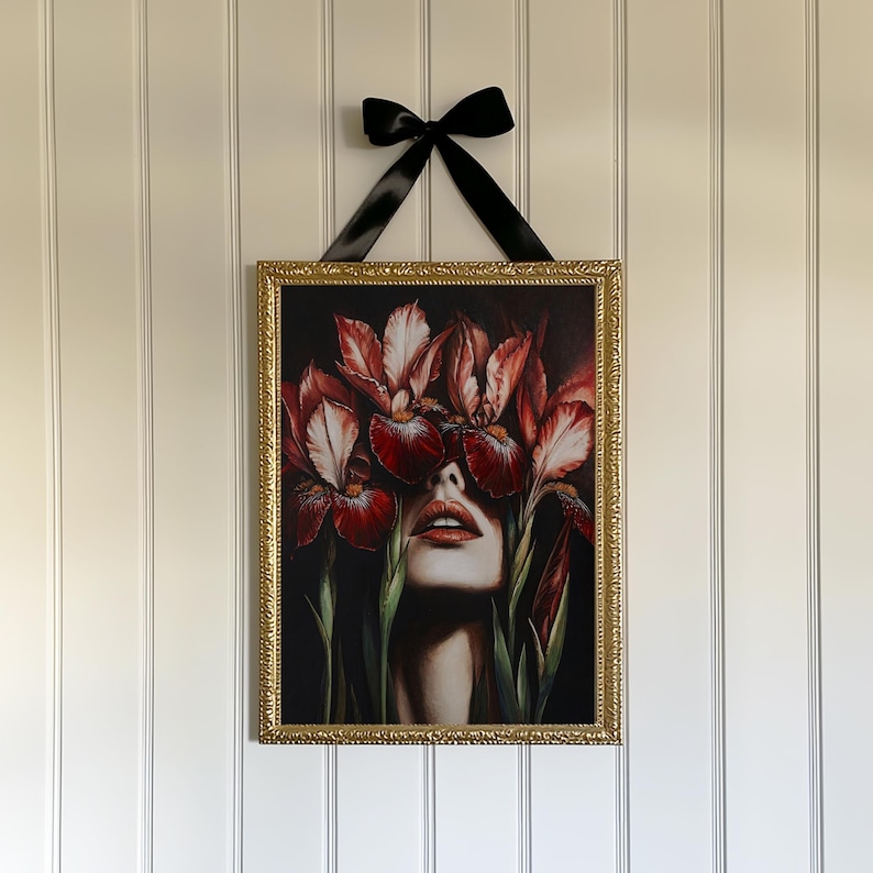 May include: A framed print of a woman's face with red and white iris flowers covering her eyes. The flowers are in focus, and the woman's face is blurred. The frame is gold and ornate.