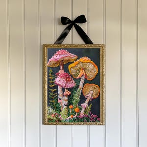 Puede incluir: Una obra de bordado enmarcada que presenta un grupo de setas coloridas con sombreros rosas y naranjas sobre un fondo oscuro. Las setas est&aacute;n rodeadas de follaje verde y peque&ntilde;as flores.