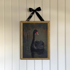 May include: A framed print of a black swan swimming in a dark pond. The frame is gold with an ornate design.