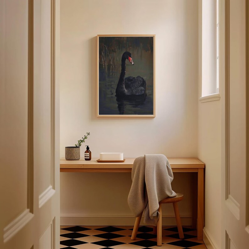 May include: A framed print of a black swan swimming in a dark green pond. The swan has a red beak and is facing to the right. The print is hanging on a white wall above a wooden desk with a plant, a bottle of lotion, and a white rectangular object.