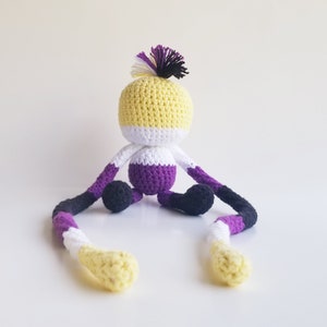 May include: A crocheted plush toy with long arms and legs. The toy is sitting with its legs crossed. The toy is yellow, white, purple, and black. The toy has a small tuft of yarn on its head.