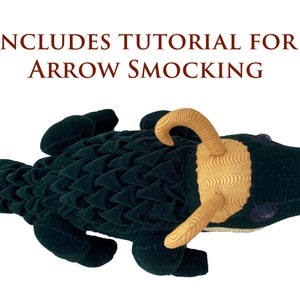 Op de afbeelding: Een pluche groene alligator speelgoed met een gouden en bruine hoorn. De rug van de alligator is bedekt met een textuur stof dat lijkt op schubben. Het speelgoed bevat een tutorial voor pijl smocking.
