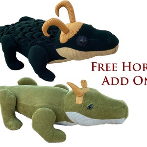 Op de afbeelding: Twee pluche alligator speelgoedjes, een groen en een zwart, met gele hoorns. De tekst "FREE HORNS ADD ON" wordt onder de speelgoedjes weergegeven.