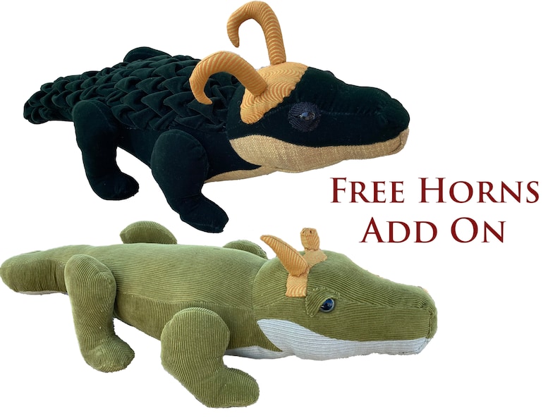 Op de afbeelding: Twee pluche alligator speelgoedjes, een groen en een zwart, met gele hoorns. De tekst "FREE HORNS ADD ON" wordt onder de speelgoedjes weergegeven.