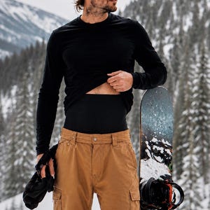 May include: A black long-sleeve shirt worn over a black compression waist band, paired with tan cargo pants. A snowboard with black bindings stands upright next to the pants. The clothing is worn in a snowy mountain environment.