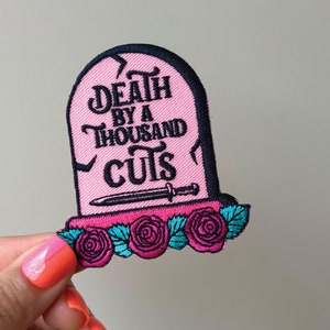 May include: Pink embroidered patch in the shape of a tombstone with the words "Death by a Thousand Cuts". The patch features a small dagger and is decorated with pink roses and teal leaves.