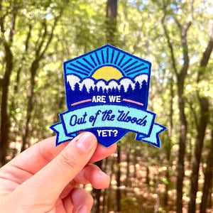 May include: An embroidered patch in blue, teal, and yellow, with the text "ARE WE Out of the Woods YET?". The design includes a mountain range, sun, and sky. The patch is held up against a blurred backdrop of trees.
