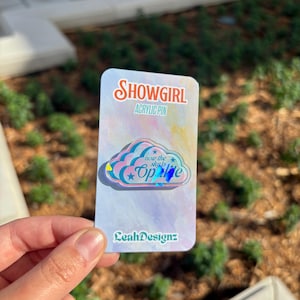 May include: An acrylic pin with the text "SHOWGIRL" and "ACRYLIC PIN" in a cursive font. The pin features a cloud design with the text "now the sky is Opale". The pin is held in a hand.