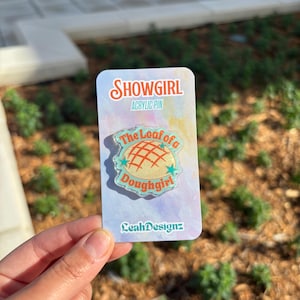 May include: An acrylic pin with the text "SHOWGIRL" and "ACRYLIC PIN" at the top. The pin features a design of a loaf of bread with the text "The Loaf of a Doughgirl". The pin is held in a hand.