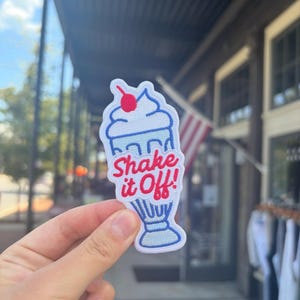 May include: Embroidered patch featuring a cartoon milkshake with a cherry on top. The patch has a white background with blue and red accents, and the text "Shake it Off!" in red. The patch is held in front of a blurred outdoor background.